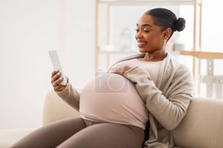 Photo for Smiling pregnant african american woman websurfing on smartphone at home, sitting on couch, watching video content, scrolling, reading blog for future moms, copy space - Royalty Free Image