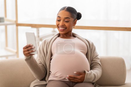 Foto de Mujer embarazada negro alegre usando teléfono inteligente mientras se relaja en el sofá en casa, señora feliz esperando bebé viendo videos divertidos y sonriendo, espacio de copia para la publicidad - Imagen libre de derechos