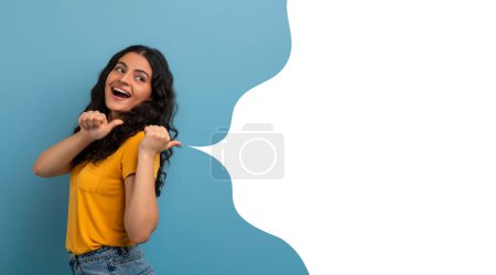 Foto de Consulta esta oferta. Joven mujer india alegre señala pulgares a la burbuja del habla, mostrando el espacio vacío de la bandera listo para el texto de marketing, sobre fondo de estudio azul, panorama, collage - Imagen libre de derechos