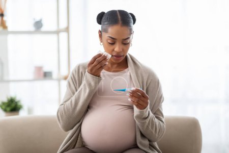 Flu during pregnancy. Unhealthy young african american pregnant woman checking body temperature, sitting on couch at home, holding thermometer and napkin, suffering from cold, copy space
