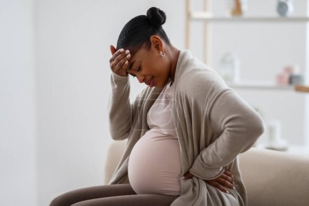 Téléchargez les photos : Migraine pendant la grossesse. Afro-américain attend femme souffrant de maux de tête à la maison, assis sur le canapé dans le salon confortable, toucher la tête, se sentir mal, copier l'espace - en image libre de droit