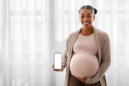 Téléchargez les photos : Joyeux jeune femme noire enceinte montrant téléphone portable avec écran blanc blanc maquette copie espace, debout à côté de la fenêtre à la maison, embrasser le ventre, application pour les futures mamans - en image libre de droit