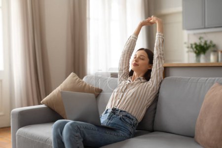 Téléchargez les photos : Contentée jeune femme en chemisier rayé étire les bras au-dessus, profitant d'une pause tout en travaillant sur un ordinateur portable, assise sur un canapé gris, espace libre - en image libre de droit