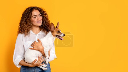 Téléchargez les photos : Jeune femme aux cheveux bouclés berce doucement son heureux Jack Russell Terrier, à la fois profiter du moment, sur fond jaune, symbolisant la joie et la compagnie - en image libre de droit