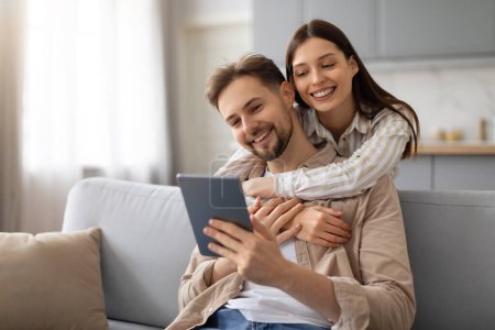 Photo for Cheerful young couple sharing cozy moment on sofa, woman hugging man from behind, they both engage with digital tablet, having video call or surfing internet - Royalty Free Image