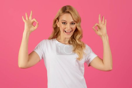 Téléchargez les photos : Jeune femme blonde souriante montrant OKAY geste avec les deux mains sur fond de studio rose. Joyeux millénium dame donnant un retour positif et exprimant son approbation - en image libre de droit