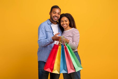 Photo for Cheerful young black couple holding bright paper shopper bags and smiling at camera, happy african american spouses enjoying day out shopping, standing on yellow studio background, copy space - Royalty Free Image