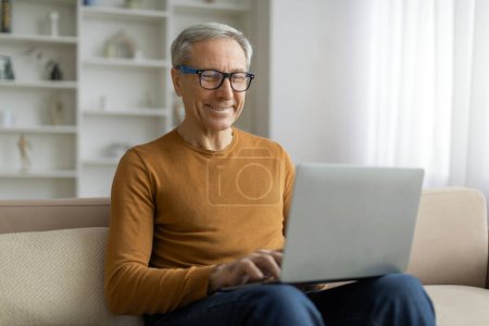 Positive senior man wearing eyeglasses using laptop at home, sitting on couch with computer on his lap, websurfing, reading news online, banking on Internet. Digital world and elderly people