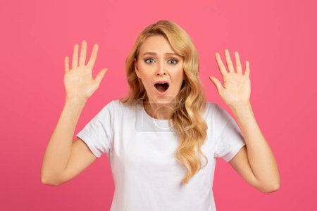 Photo for Shocked emotional blonde woman got caught posing with raised arms looking at camera with amazed upset expression, standing over pink studio background. Omg, its not me concept - Royalty Free Image