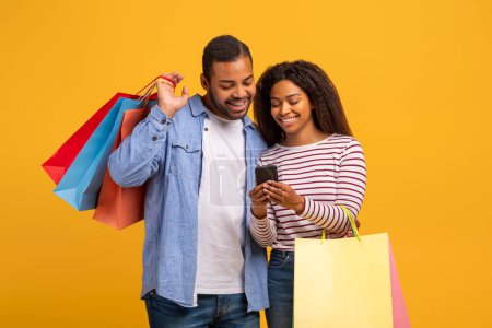 Photo for Shopping App. African American Couple Holding Shopper Bags And Using Smartphone, Happy Young Black Spouses Browsing Mobile Application With Sale Offers And Discounts, Standing Over Yellow Background - Royalty Free Image