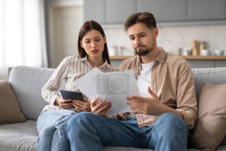 Photo for Concerned young spouses sitting on gray sofa, attentively examining papers and using digital tablet, possibly discussing finances or plans in their living room - Royalty Free Image