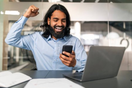 Photo for Excited happy millennial eastern businessman in formal outwear sitting at modern office, looking at phone screen, gesturing and exclaiming, celebrating good news, business success, copy space - Royalty Free Image