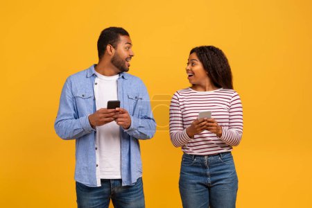 Téléchargez les photos : Portrait de joyeux jeune couple noir tenant des smartphones et se regardant les uns les autres, heureux Afro-Américain homme et femme utilisant des téléphones cellulaires tout en se tenant ensemble sur fond jaune, Copier l'espace - en image libre de droit