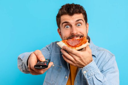 Téléchargez les photos : Émotionnel jeune homme européen aime pizza tranche pointant télécommande à la caméra, regarder l'émission de télévision préférée et la frénésie de manger de la malbouffe, sur fond de studio bleu, gros plan portrait - en image libre de droit