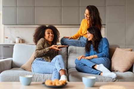 Foto de Trío multicultural de señoritas charlando alegremente alrededor de la mesa de la sala de estar, compartiendo galletas y café en un ambiente relajado en casa, disfrutando de la conversación, el tiempo juntos y la amistad - Imagen libre de derechos