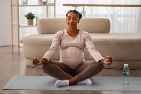 Photo for Peaceful african american young woman sitting in lotus pose with closed eyes on yoga mat, meditating at home, copy space. Fitness during pregnancy, relaxation, stress relief - Royalty Free Image