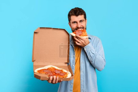 Photo for Hungry millennial man holds pizza box taking bite of tasty slice on blue studio background, showcasing concept of cheat meal, overeating habit and enjoying taste of junk food - Royalty Free Image