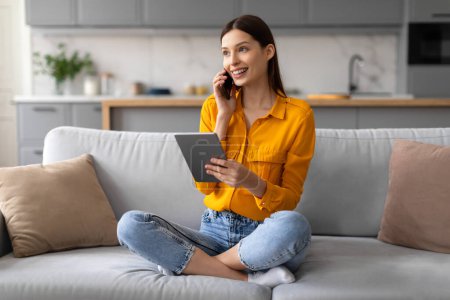Cheerful young woman comfortably sitting on sofa, simultaneously engaging with digital tablet and talking on smartphone, managing her tasks with ease at home