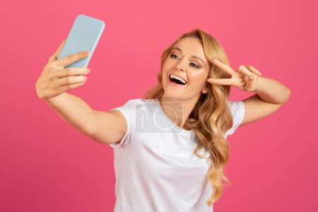 Photo for Cheerful blonde young woman in casual making selfie on smartphone, gesturing victory sign, posing on pink studio backdrop, having fun online while making content for social media - Royalty Free Image
