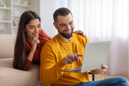 Foto de Feliz pareja joven websurf en el ordenador portátil juntos mientras se relaja en casa, alegres cónyuges milenarios de compras en línea, descansando con el ordenador en la sala de estar, hombre apuntando a la pantalla, espacio de copia - Imagen libre de derechos