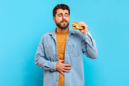 Téléchargez les photos : Manger malsain. Le jeune homme touche son estomac endolori tenant un hamburger en studio sur fond bleu, ressentant de l'inconfort après une consommation de malbouffe. Gastrite, mauvais choix alimentaires - en image libre de droit