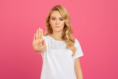 Téléchargez les photos : Déterminé jeune femme blonde geste arrêter avec la main, montrant signe de rejet et de protestation contre fond de studio rose, portrait tourné. Halte aux abus, à la violence et à la discrimination - en image libre de droit
