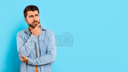 Portrait of thoughtful millennial man in casual outfit, captured deep in contemplation, thinking while touching chin against blue studio background, looking aside at copy space, panorama