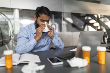 Foto de Unwell millennial empresario indio en camisa azul sonándose la nariz, con medicina en el escritorio, indicando síntomas de resfriado o gripe mientras trabaja, interior moderno de la oficina, espacio para copiar - Imagen libre de derechos