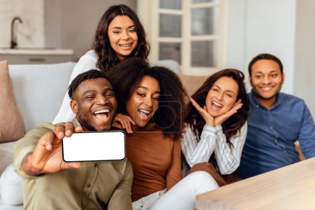 Photo for Joyful group of young multiethnic friends making selfie using mobile phone with empty screen, capturing joyful moment of friendship and celebration, posing together in living room indoors - Royalty Free Image