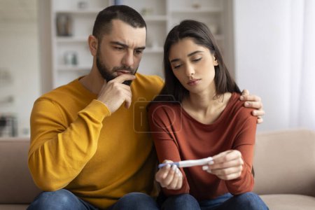 Photo for Infertility Problems. Portrait Of Upset Young Couple Looking At Negative Pregnancy Test While Sitting Together On Couch At Home, Caring Husband Embracing Depressed Wife, Free Space - Royalty Free Image