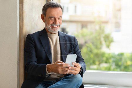Photo for Smiling european older man using a smartphone by the window, work in office, chatting and typing message, read news. Meeting and communication, business remotely, video call - Royalty Free Image