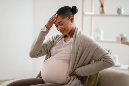 Photo for African american pregnant woman suffering from headache at home, sitting on couch in cozy living room, touching her head, feeling unwell, migraine during pregnancy, copy space - Royalty Free Image