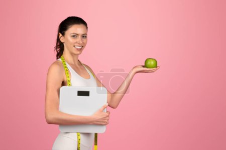 Téléchargez les photos : Femme entraîneur d'ajustement caucasienne soucieuse de sa santé avec un ruban à mesurer autour du cou tenant une pomme verte et une balance, symbolisant une nutrition équilibrée et la gestion du poids - en image libre de droit