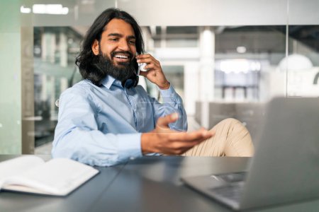 Photo for Corporate Communication. Smiling Indian Guy Talking On Cellphone Working On Computer At Office, Positive Guy Sitting At Desk With Laptop Having Phone Conversation, Enjoying Job, Free Copy Space - Royalty Free Image