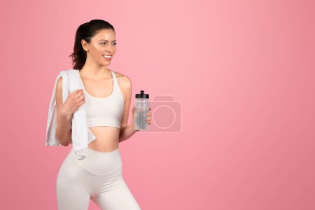 Photo for A smiling caucasian fitness enthusiast in white attire carries a water bottle and a towel over her shoulder, ready for a hydration break during her active workout routine - Royalty Free Image