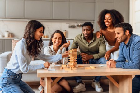 Foto de Grupo de alegres amigos diversos millennials se reúnen alrededor de la mesa en la sala de estar, participar en un animado juego de mesa, la construcción de una torre de madera de bloques y divertirse juntos el fin de semana - Imagen libre de derechos