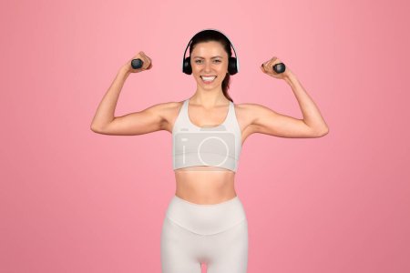 Photo for Joyful european woman with a beaming smile flexing her muscles wearing headphones and a white sports bra and leggings, embodying a fun and active fitness lifestyle on a pink background - Royalty Free Image