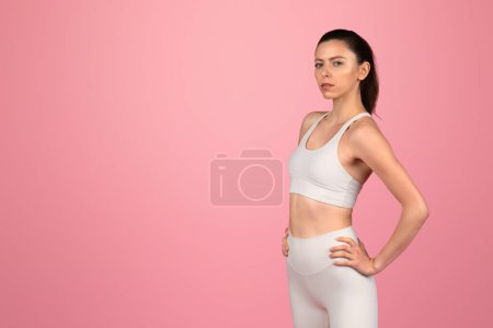 Foto de Decidida joven europea confiada en traje de fitness blanco de pie con confianza con las manos en las caderas, mostrando fuerza y determinación, contra un fondo rosa, estudio - Imagen libre de derechos