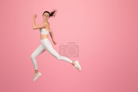 Photo for Energetic european young woman in motion, joyfully running in mid-air with a white sports bra and leggings against a pink backdrop, portraying vitality and an active lifestyle - Royalty Free Image