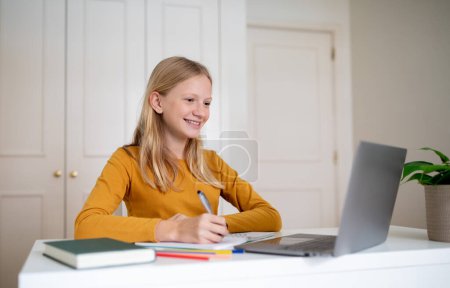 Foto de Concepto de Educación en Línea. Sonriendo joven adolescente usando el ordenador portátil y tomando notas mientras está sentado en el escritorio en casa, feliz adolescente niña mirando a la pantalla del ordenador portátil, haciendo la tarea, espacio libre - Imagen libre de derechos