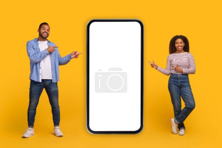 Photo for Place For Ad. Cheerful African American Couple Demonstrating Big Blank Smartphone With White Screen For Mockup And Pointing At It, Happy Black Man And Woman Advertising New Mobile App Or Website - Royalty Free Image