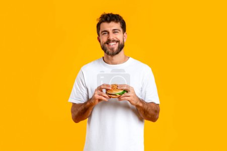 Téléchargez les photos : Heureux homme européen en t-shirt blanc posant tenant un cheeseburger sur fond jaune, l'expérience de la joie de la malbouffe trop manger. Bannière pour l'offre de restauration rapide, concept de repas de triche - en image libre de droit