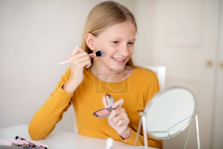 Photo for Smiling teen girl applying blush with makeup brush at home, happy cute teenage female child learning to do make up, sitting at desk in room and looking in mirror, closeup shot with free space - Royalty Free Image