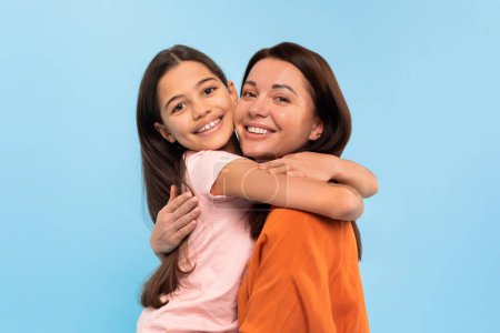 Téléchargez les photos : Portrait de sourire jeune mère et mignonne Preteen fille embrassant tout en posant sur fond bleu studio, en regardant la caméra. Petite fille étreint sa maman coller ensemble - en image libre de droit