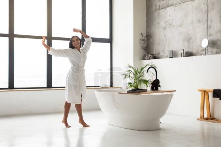 Foto de Mujer india joven y despreocupada que usa albornoz blanco bailando en un baño de lujo con bañera y ventana panorámica, longitud completa, espacio para copiar. Feliz señora oriental disfrutando de buenos días - Imagen libre de derechos
