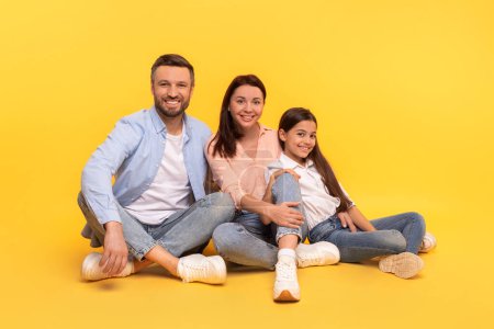 Téléchargez les photos : Famille aimante est assis de près posant sur fond jaune, plan studio. Mère, père et fille heureux ensemble, assis et souriant chaleureusement, exprimant le bonheur et le lien familial - en image libre de droit