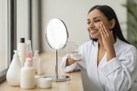 Skin care routine. Happy attractive young indian woman in white bathrobe using face cream at home, beautiful lady test newest organic beauty product, sitting at vanity table, copy space