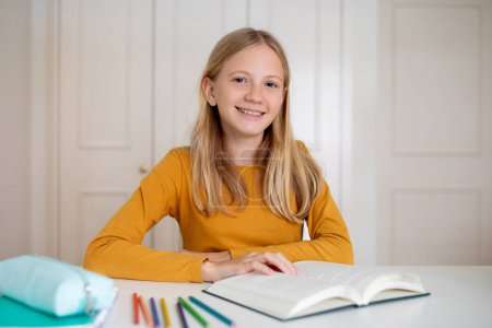 Téléchargez les photos : Adolescente souriante assise au bureau avec un livre ouvert, des marqueurs colorés et un étui à crayons à la maison, heureuse jeune adolescente mignonne prête à étudier, appréciant la lecture et l'éducation, espace de copie - en image libre de droit