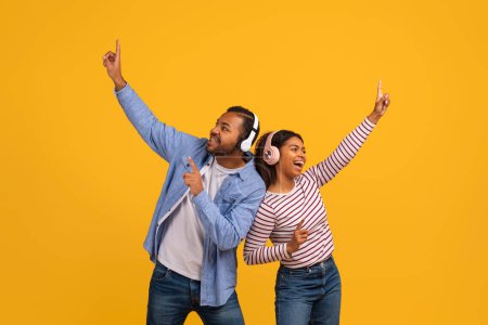 Téléchargez les photos : Joyeux jeune couple noir portant des écouteurs jouissant de musique et dansant ensemble, homme et femme afro-américain joyeux s'amuser, debout isolé sur un fond jaune vibrant, espace de copie - en image libre de droit