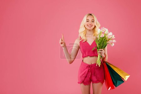 Photo for Cheerful blonde woman with bouquet of tulips and colorful shopping bags, gesturing an idea on pink background, celebrating Womens Day, pointing at free space - Royalty Free Image